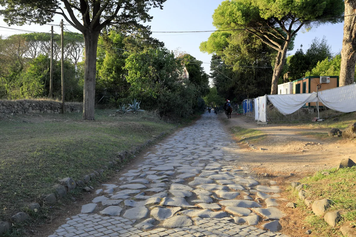 A stretch of the Appian Way paved with large irregular stone slabs between grass verges and umbrella pines
