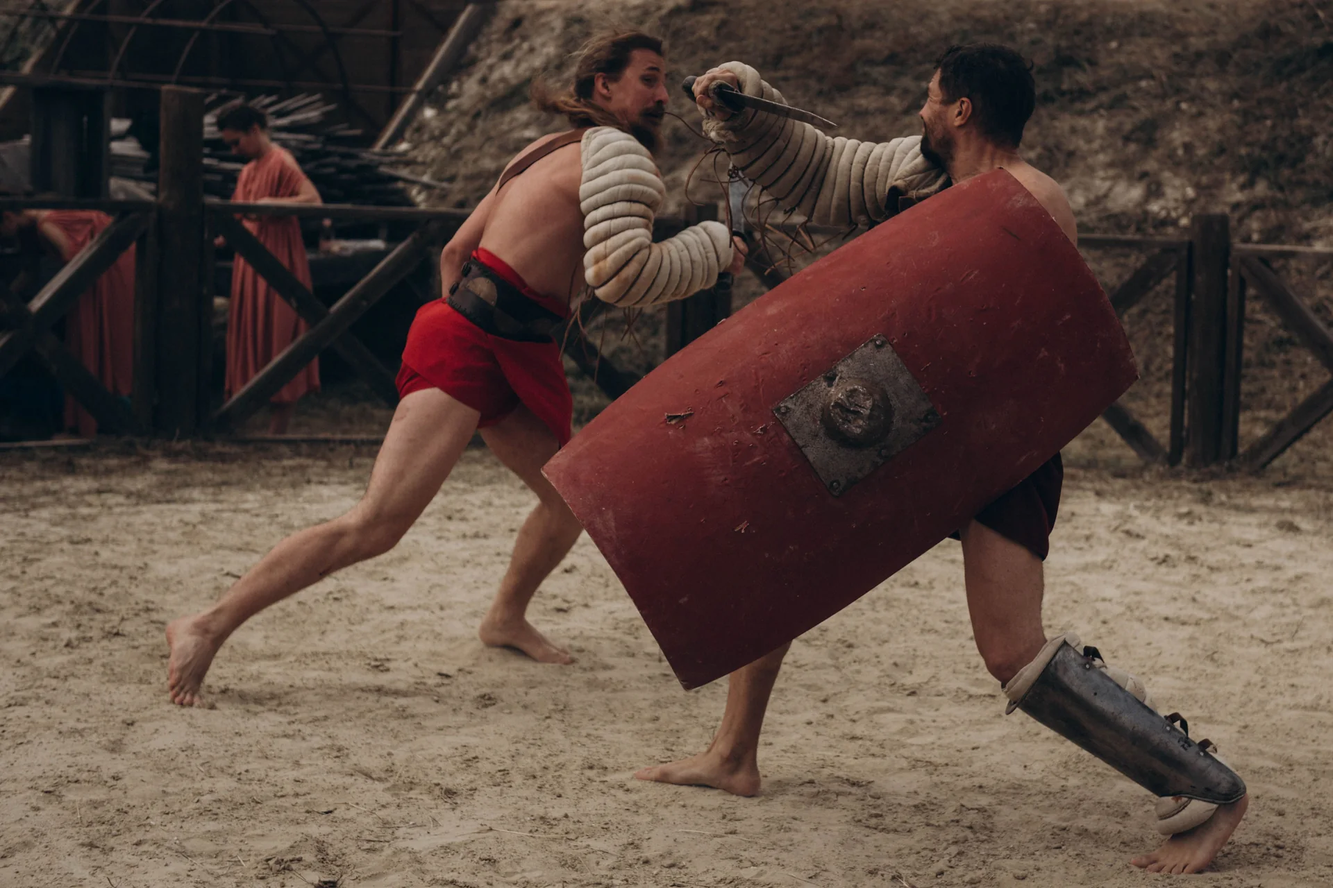 Two gladiator re-enactors fight in a sand arena, one lunging with a dagger over a large red shield while the other, bare-chested in a red tunic and padded arm guard, twists away