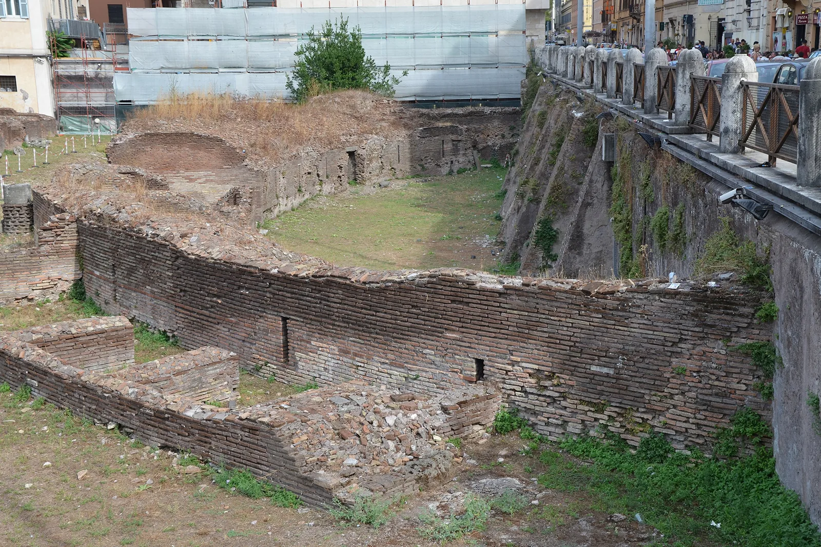 The ruins of the Ludus Magnus beside the Colosseum, an oval of brick walls around a sunken space with a road running alongside