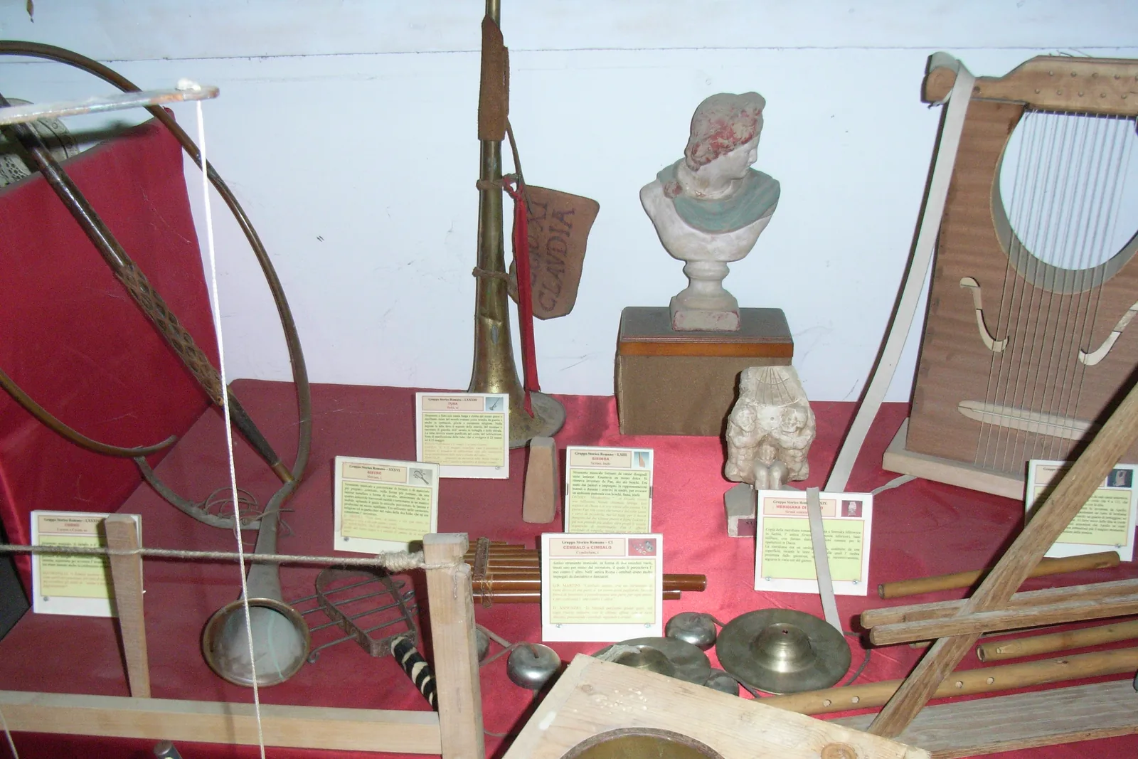 A museum display of reproduction Roman musical instruments, including a bronze horn, a lyre, pan pipes and cymbals, with a plaster bust and printed labels on a red cloth